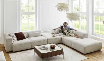 Großzügiges, helles Wohnzimmer mit bodentiefen Fenstern und modernem, beigem Ecksofa. Ein Paar sitzt entspannt auf dem Sofa und genießt gemeinsam ein Buch.